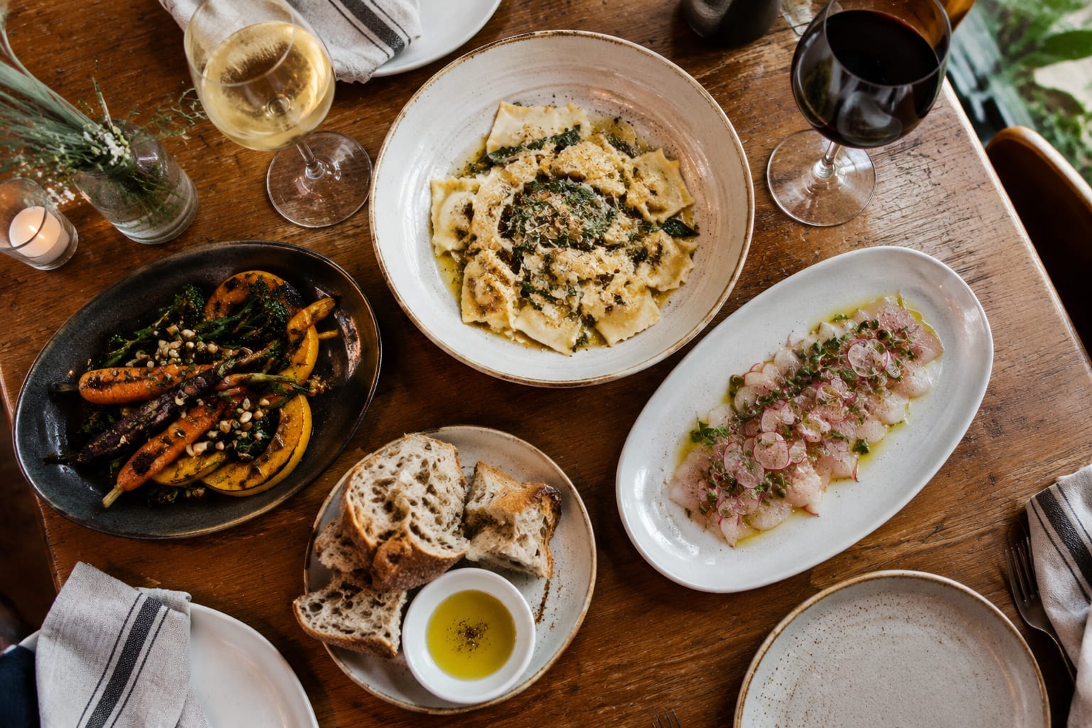 Overhead view of shared plates of pasta, crudo, roast vegetables and wine on a restaurant table