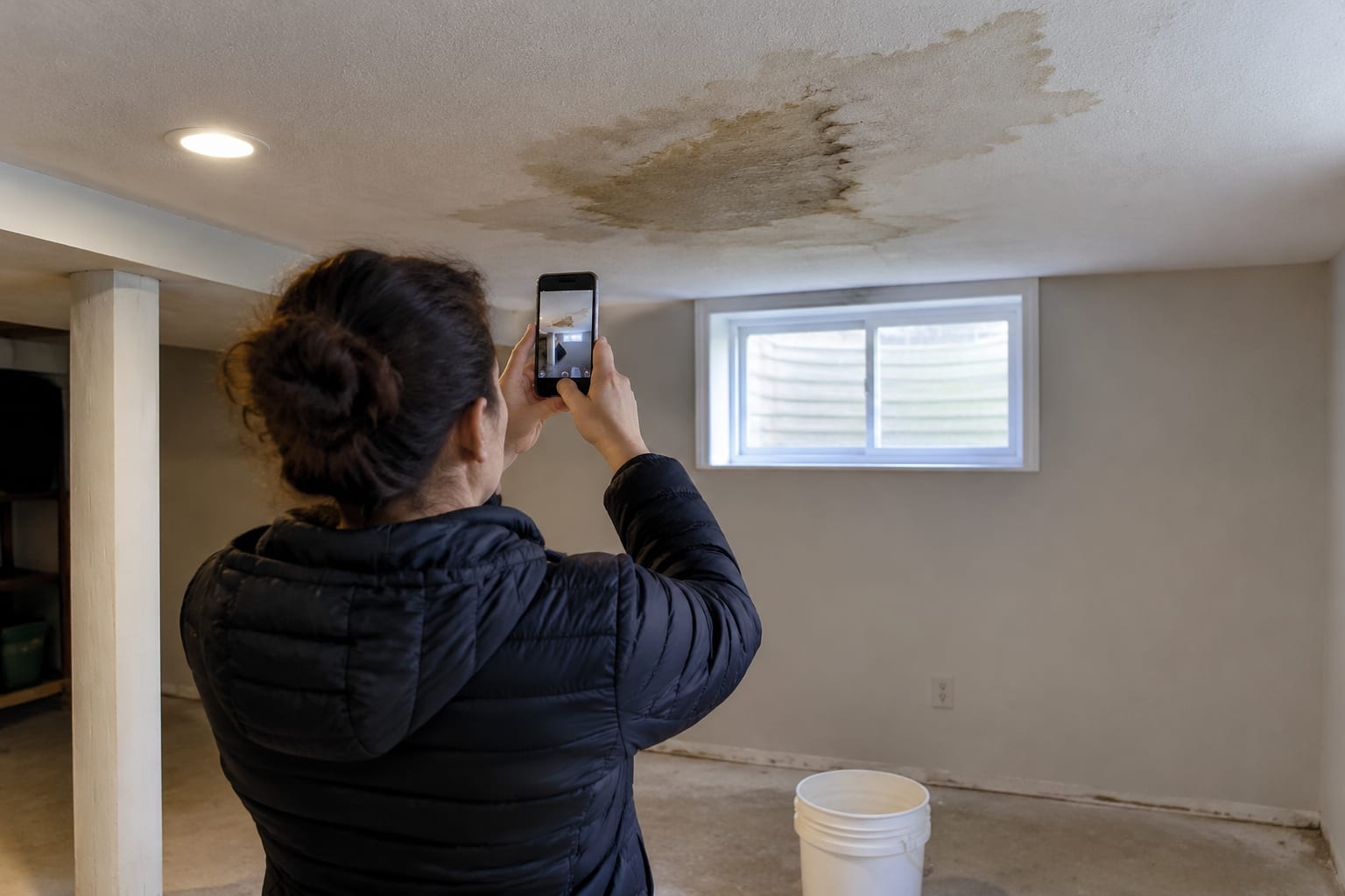 A homeowner on the phone photographing a water-stained basement ceiling with a smartphone