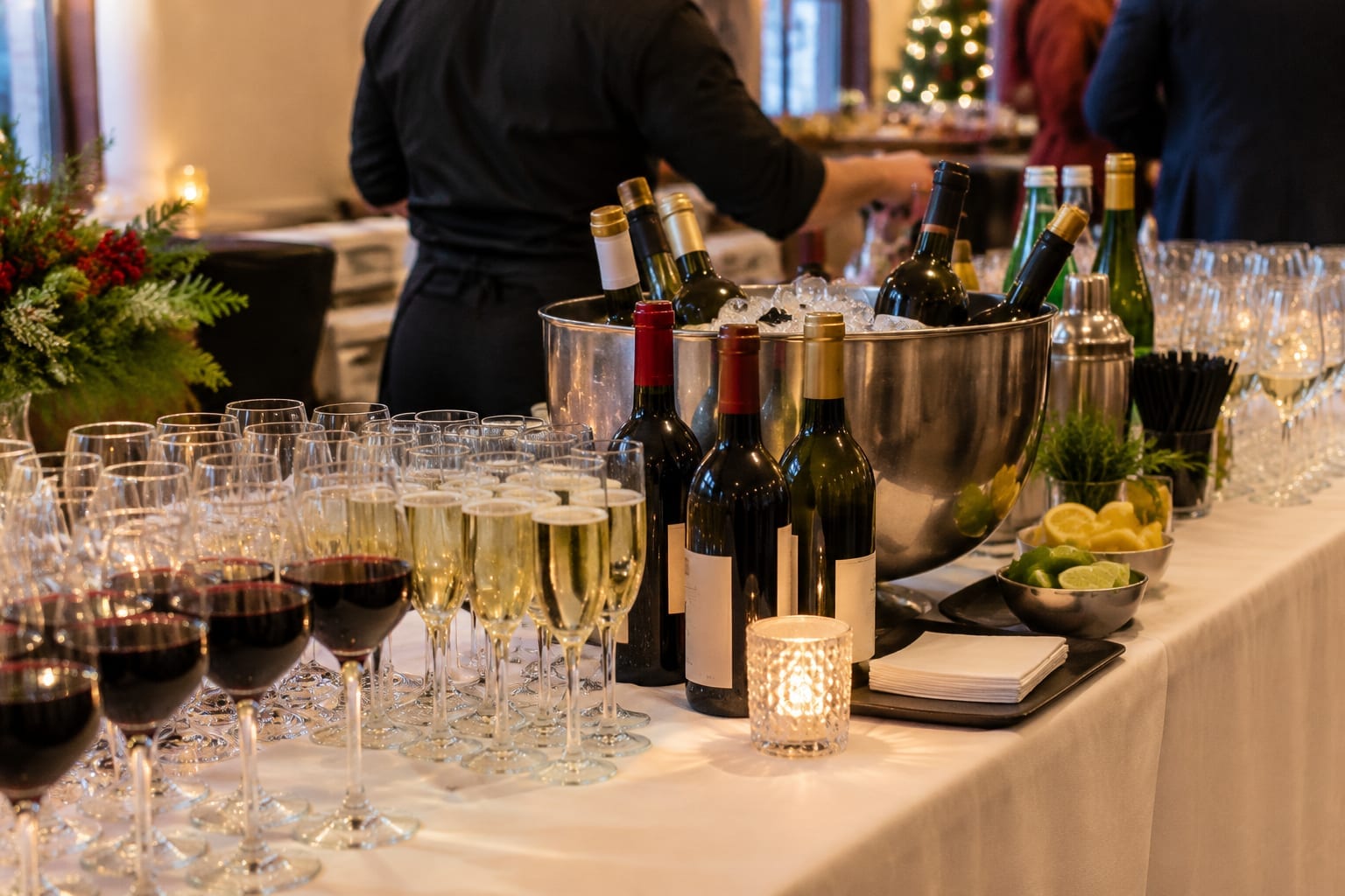 A small bar setup with glasses, wine bottles and a bartender in black preparing drinks at a party