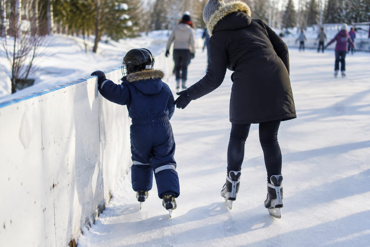 A child in a snowsuit and hockey helmet holding the boards at an outdoor rink while a parent skates beside them