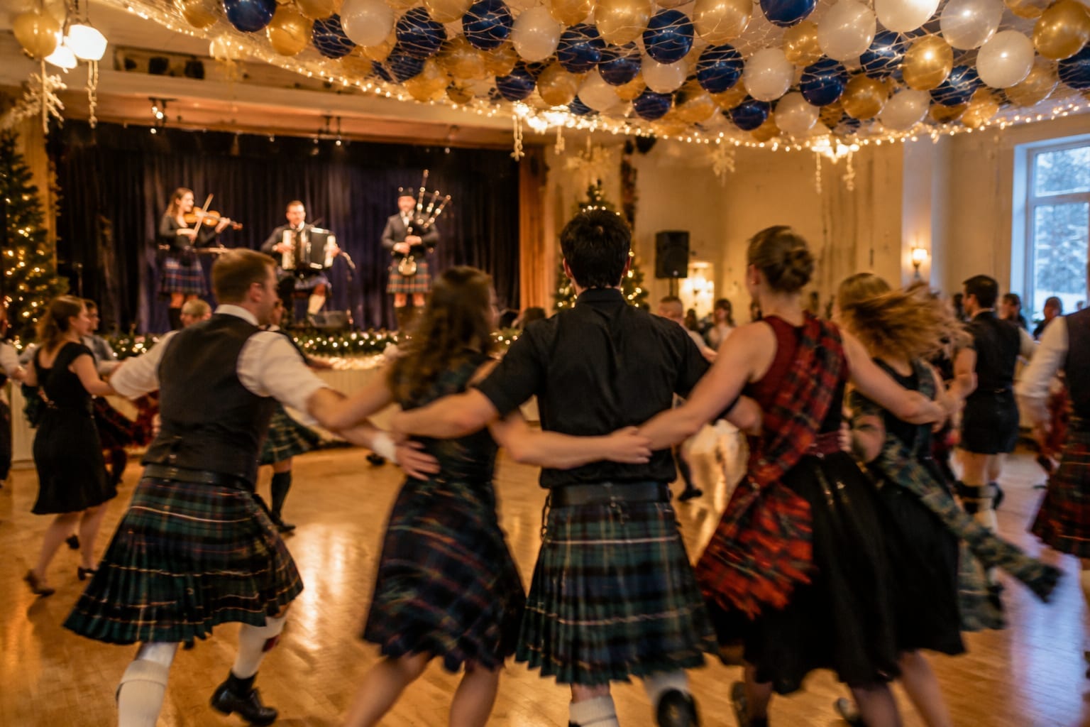 Ceilidh band with fiddle and bagpipes as kilted dancers link arms in a hall