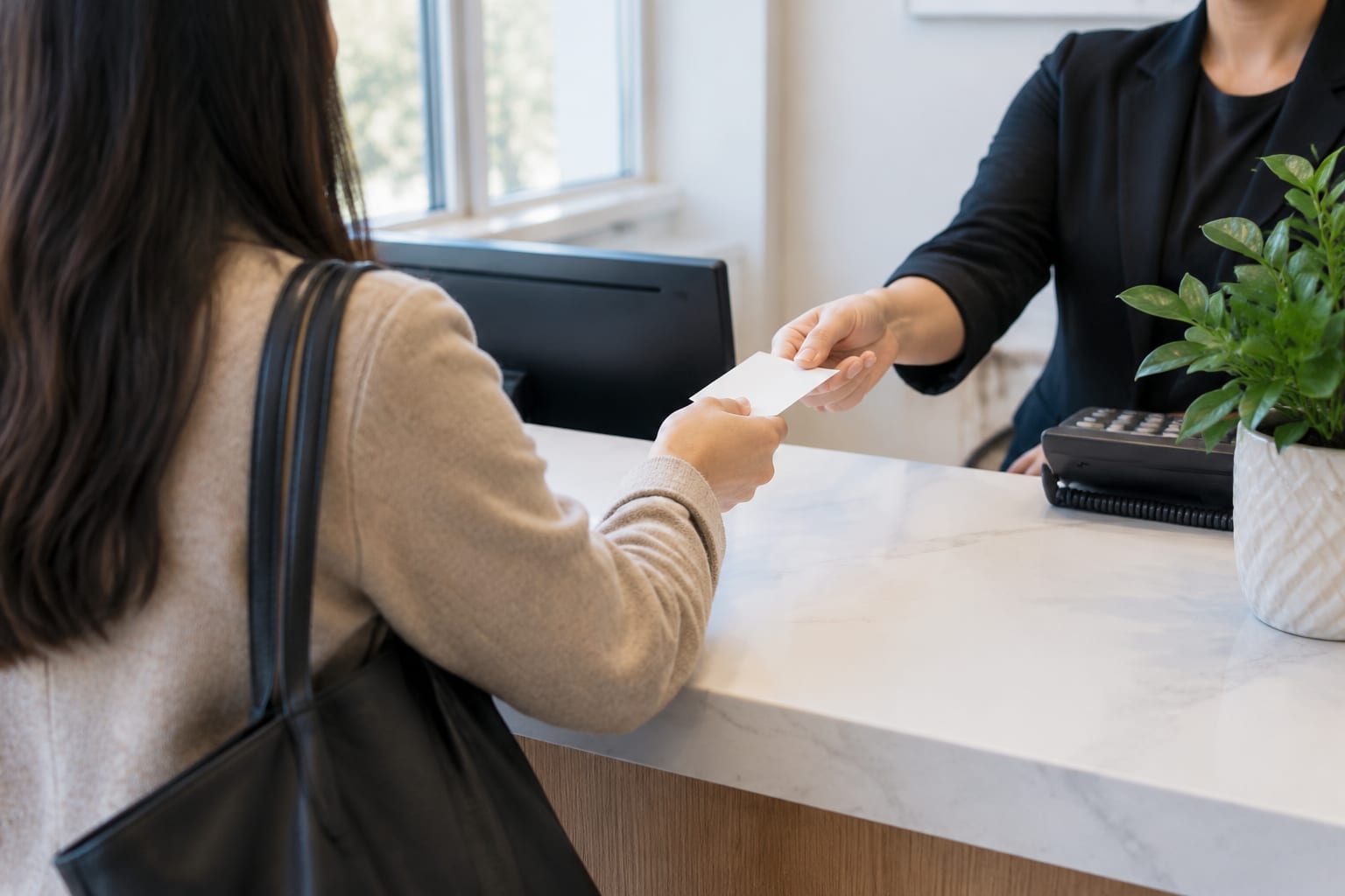 A person at a clinic reception desk handing a plain card to a receptionist, seen from behind